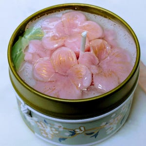 May include: A floral-themed candle in a decorative tin. The candle features pink flower petals with gold accents, set in a clear wax. The tin is green with a floral pattern and a gold rim. A white wick is centered in the candle.