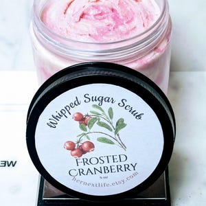 May include: A jar of Frosted Cranberry Whipped Sugar Scrub, with a pink, fluffy texture. The black lid features a label with cranberry illustrations and the text "Whipped Sugar Scrub" and "Frosted Cranberry". The jar is on a scale that reads 12.13 oz.