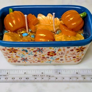 May include: A decorative ceramic baking dish with a blue rim and floral pattern, filled with orange pumpkin-shaped candles and other wax elements. The dish is approximately 6 inches long, and includes a ruler for scale.