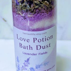May include: A clear glass bottle with a silver lid, filled with layers of bath dust. The label reads "Love Potion Bath Dust Lavender Fields" with the Etsy shop URL. The bottle contains dried lavender and other ingredients.