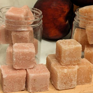 May include: Pink, cube-shaped bath bombs are stacked in and out of clear glass jars. The bath bombs have a slightly textured surface. A wooden cutting board is in the foreground. Peaches are in the background.