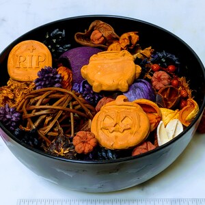 May include: A black bowl filled with Halloween-themed potpourri, including orange clay shapes of a tombstone with RIP text, a cauldron, and a jack-o'-lantern, mixed with purple, brown, and orange dried botanicals and a woven twig sphere.