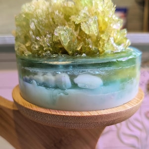 May include: A decorative, layered soap with a light green, blue, and white color scheme. The top of the soap features a cluster of light green, crystal-like formations. The soap is displayed on a round, wooden stand.
