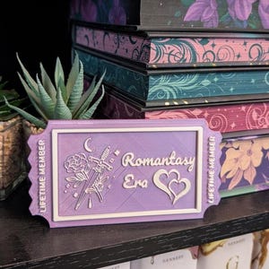 May include: A lavender "Romantasy Era" lifetime member ticket featuring a rose, sword, and heart design. The ticket sits on a shelf with a stack of floral-decorated books and a small succulent plant.