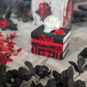 Puede incluir: Una pila de tres libros decorativos con portadas negras y rojas, que presentan el texto "Dark ROMANCE" en relieve. Una calavera blanca se sitúa sobre la pila, acompañada de pequeñas rosas rojas en los lomos y la portada.