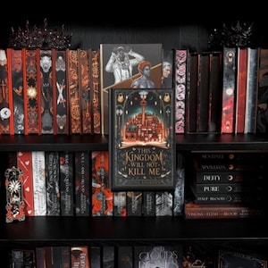 Op de afbeelding: Een verzameling boeken op zwarte planken, met een centraal hardcover boek getiteld This Kingdom Will Not Kill Me van Ilona Andrews, omringd door diverse fantasyromans met decoratieve ruggen, donkere covers en twee zwarte kronen.