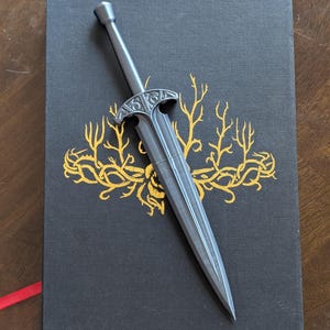 May include: A silver-colored letter opener in the shape of a sword rests on a black book with gold floral designs. The book has a red ribbon bookmark. The letter opener has a detailed hilt.