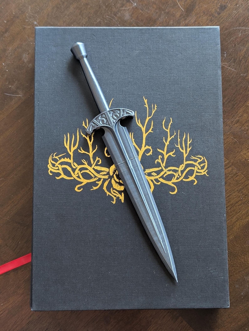 May include: A silver-colored letter opener in the shape of a sword rests on a black book with gold floral designs. The book has a red ribbon bookmark. The letter opener has a detailed hilt.