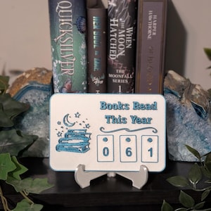 May include: A white and blue wooden sign displaying "Books Read This Year" and the number "061". The sign is supported by a silver stand, with several books visible in the background.