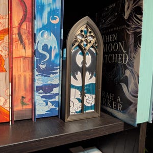 Könnte beinhalten: Ein dekoratives Fenster im gotischen Stil mit Fledermaus-Design, ausgestellt auf einem Bücherregal. Das Fenster ist dunkelbraun gerahmt und hat ein detailliertes, durchbrochenes Design an der Oberseite. Der Hintergrund zeigt ein türkis-weißes Muster.