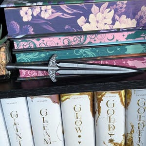 May include: A decorative silver letter opener with an ornate handle rests on a stack of books. The books have floral and celestial designs in shades of purple, pink, and teal. The spines of the books below read "Glint", "Gleam", "Glow", and "Gold".