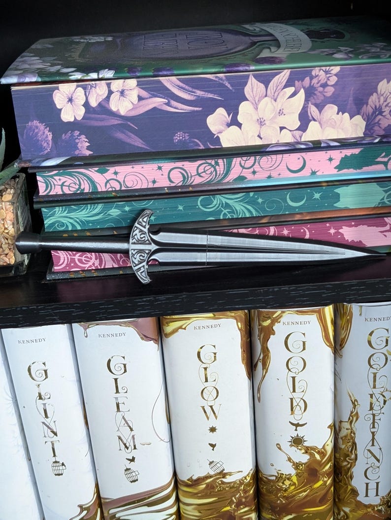 May include: A decorative silver letter opener with an ornate handle rests on a stack of books. The books have floral and celestial designs in shades of purple, pink, and teal. The spines of the books below read "Glint", "Gleam", "Glow", and "Gold".