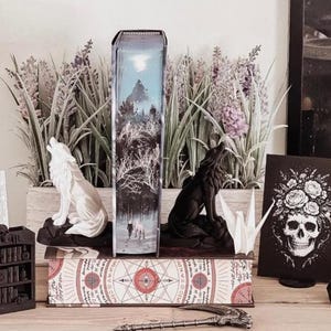 May include: A set of two wolf bookends, one white and one black, flanking a book with a winter forest scene on the spine. The book rests on a base featuring red circular geometric patterns and text. A small metal scythe lies in the foreground.
