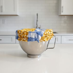 May include: A stainless steel mixing bowl with a handle is topped with a fabric bowl cover. The cover features a yellow floral pattern with black accents and a blue and white gingham pattern. The kitchen setting includes white cabinets and a countertop.