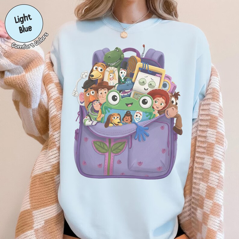 Puede incluir: Una sudadera de cuello redondo azul claro con un estampado gr&aacute;fico de una mochila morada llena de varios personajes de juguetes animados, incluyendo una rana verde, un vaquero, un guardi&aacute;n espacial y una vaquera, en un estilo de dibujo animado.