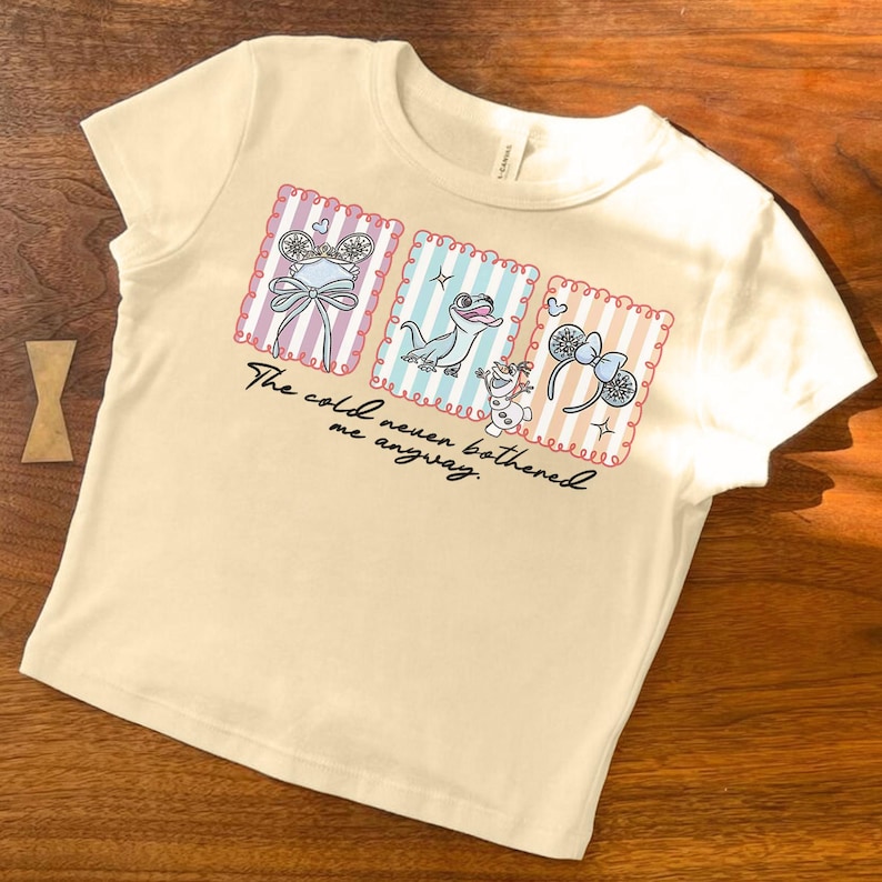 May include: A cream-coloured t-shirt with three framed illustrations. The illustrations feature a bow, a lizard, and a snowman. The text "The cold never bothered me anyway" is printed below the illustrations. The t-shirt is made of a soft material.