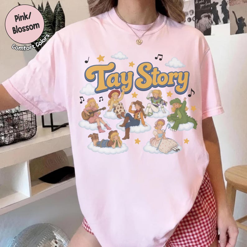 May include: A light pink short-sleeve t-shirt featuring the yellow text Tay Story. The graphic includes multiple illustrated characters sitting on clouds, surrounded by small yellow stars, musical notes, and white clouds on a light pink background.