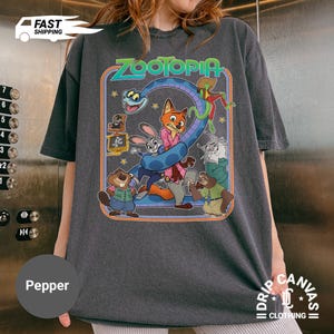 May include: Dark gray t-shirt featuring a colorful graphic from the movie Zootopia. The design includes various animal characters and the word "ZOOTOPIA" in a retro style. The shirt has a relaxed fit and short sleeves.