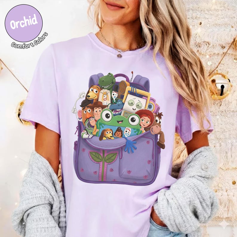 Puede incluir: Una camiseta de color morado claro con un estampado gr&aacute;fico de una mochila morada llena de varios personajes de dibujos animados, incluyendo una rana verde, un vaquero, un ranger espacial y otras figuras animadas.