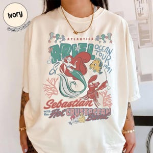 May include: An ivory t-shirt featuring a graphic print of Ariel, Flounder, and Sebastian. The text reads Atlantica Ariel Ocean Tour 1991, Sebastian with the Hot Crustacean Band. The design includes coral, bubbles, and sea-themed illustrations in muted tones.