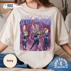 May include: Ivory-colored t-shirt featuring a graphic of Judy Hopps from Zootopia. The design includes three cartoon images of Judy Hopps in a police uniform, set against a purple lightning background. The text "JUDY HOPPS" is above the images.