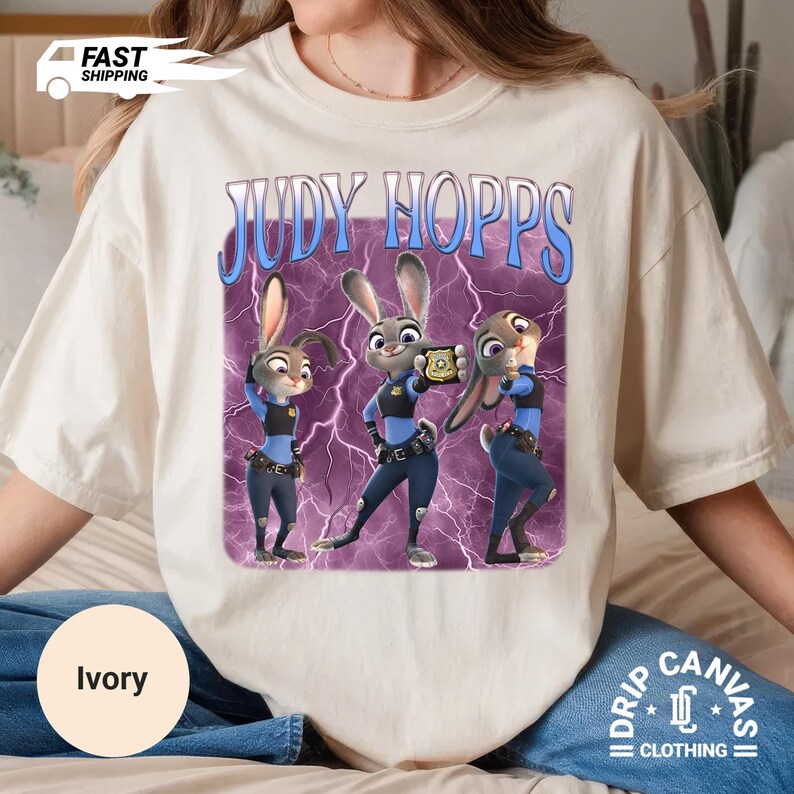 May include: Ivory-colored t-shirt featuring a graphic of Judy Hopps from Zootopia. The design includes three cartoon images of Judy Hopps in a police uniform, set against a purple lightning background. The text "JUDY HOPPS" is above the images.