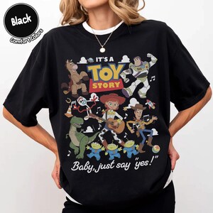 Op de afbeelding: Een zwart t-shirt met een kleurrijke afbeelding van Toy Story-personages, waaronder Woody, Buzz Lightyear, Jessie, Rex, Bullseye, Forky en de Aliens. Het ontwerp bevat de tekst "IT'S A TOY STORY" en "Baby, just say "yes!"" in wit en geel.