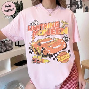 Op de afbeelding: Lichtroze T-shirt met een afbeelding van Lightning McQueen uit de film Cars. Het ontwerp bevat de tekst "Lightning McQueen" en "95", een geblokte vlag en het woord "Rusteze".