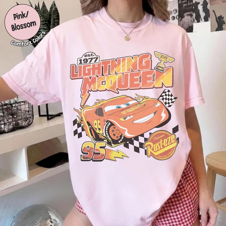 Op de afbeelding: Lichtroze T-shirt met een afbeelding van Lightning McQueen uit de film Cars. Het ontwerp bevat de tekst "Lightning McQueen" en "95", een geblokte vlag en het woord "Rusteze".