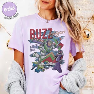 Może przedstawiać: Jasnofioletowy t-shirt z nadrukiem graficznym przedstawiającym Buzza Astrala, Zurga i zielonych kosmit&oacute;w. Tekst głosi "BUZZ Lightyear" oraz "LASER BLAST". Okrągła odznaka w lewym g&oacute;rnym rogu zawiera napis "Orchid Comfort Colors".