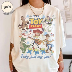 Op de afbeelding: Een ivoorkleurig t-shirt met een kleurrijke grafische print van Toy Story-personages, waaronder Woody, Buzz Lightyear, Jessie, Rex, Bullseye, Forky en drie aliens. De tekst luidt IT'S A TOY STORY en Baby, just say 'yes!' in blauw en rood.