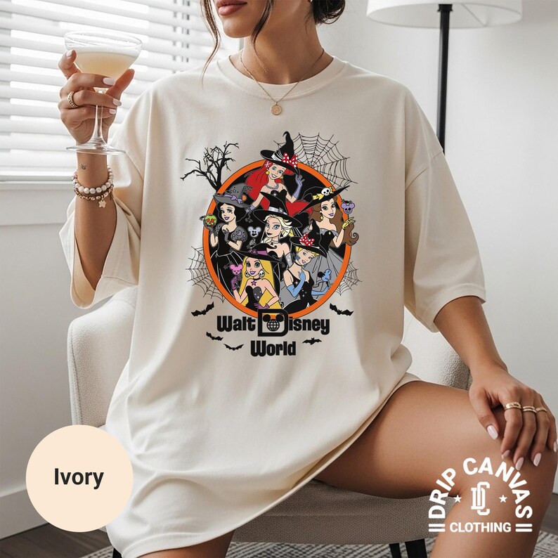 Puede incluir: Camiseta de color marfil con un dise&ntilde;o de princesas de Disney vestidas de brujas, rodeadas de una telara&ntilde;a y murci&eacute;lagos. El texto "Walt Disney World" est&aacute; impreso debajo. La camiseta es de Drip Canvas Clothing.