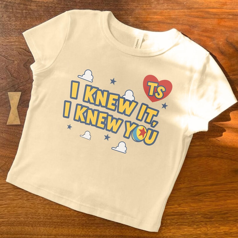 May include: A cream-colored short-sleeve t-shirt featuring the text "I KNEW IT, I KNEW YOU" in yellow with blue outlines. The design includes a red heart with "TS" inside, small blue stars, white cloud outlines, and a colorful ball graphic.