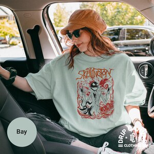 Puede incluir: Camiseta verde claro con un dise&ntilde;o rojo y negro de Jack Skellington. El dise&ntilde;o incluye el texto "Jack Skellington". La camiseta es parte de una colecci&oacute;n de ropa. La persona lleva un sombrero marr&oacute;n y gafas de sol.