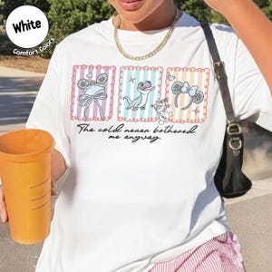 May include: White t-shirt with a colourful graphic featuring cartoon characters and the text "The cold never bothered me anyway." The design includes images of a bow, a lizard, and mouse ears. The shirt has a round neckline and short sleeves.