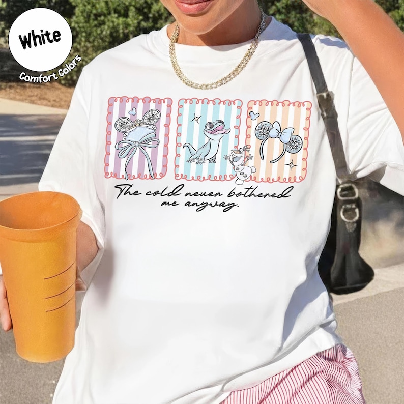 May include: White t-shirt with a colourful graphic featuring cartoon characters and the text "The cold never bothered me anyway." The design includes images of a bow, a lizard, and mouse ears. The shirt has a round neckline and short sleeves.