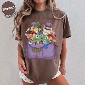 Puede incluir: Una camiseta marr&oacute;n con un gr&aacute;fico de una mochila morada llena de varios personajes de juguetes animados, incluyendo una rana verde, un vaquero, un guardi&aacute;n espacial y una vaquera. El texto Espresso Comfort Colors aparece en la esquina superior izquierda.
