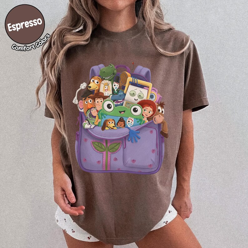 Puede incluir: Una camiseta marr&oacute;n con un gr&aacute;fico de una mochila morada llena de varios personajes de juguetes animados, incluyendo una rana verde, un vaquero, un guardi&aacute;n espacial y una vaquera. El texto Espresso Comfort Colors aparece en la esquina superior izquierda.