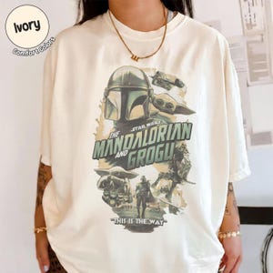 Pu&ograve; includere: T-shirt color avorio con grafica della serie Star Wars, "The Mandalorian and Grogu". Il design include immagini del Mandaloriano, Grogu e la scritta "This is the Way."