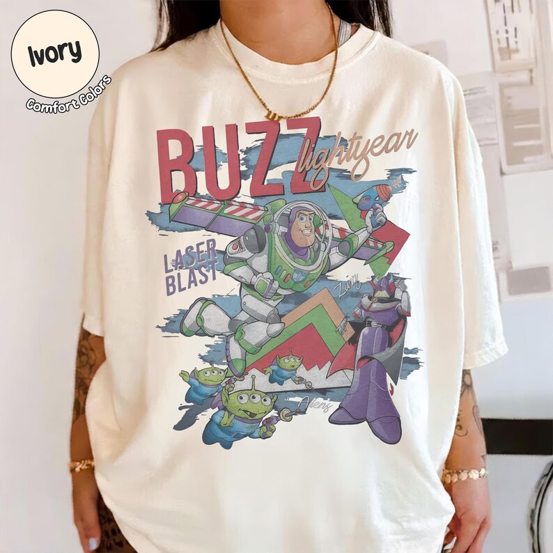 Może przedstawiać: Koszulka w kolorze kości słoniowej z nadrukiem graficznym przedstawiającym Buzza Astrala, Zurga i trzy zielone kosmity. Tekst głosi BUZZ Lightyear oraz LASER BLAST. Wz&oacute;r zawiera czerwone, zielone, fioletowe i niebieskie akcenty. Koszulka ma okrągły dekolt i kr&oacute;tkie rękawy.