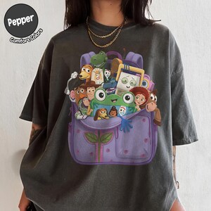 Puede incluir: Una camiseta gris carb&oacute;n con un estampado gr&aacute;fico de una mochila morada llena de varios personajes de dibujos animados, incluyendo una rana verde, un vaquero, un guardi&aacute;n espacial y un tenedor. El texto Pepper Comfort Colors aparece en la esquina superior.