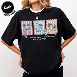 May include: Black t-shirt with three framed illustrations of characters and accessories. The text "The cold never bothered me anyway" is printed below the illustrations. The shirt has a crew neck and short sleeves.