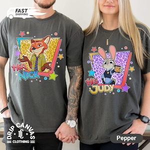 May include: Two dark gray t-shirts featuring characters from Zootopia. One shirt has Nick Wilde with "Her Nick" text, the other has Judy Hopps with "His Judy". Both shirts have colorful star and geometric designs.