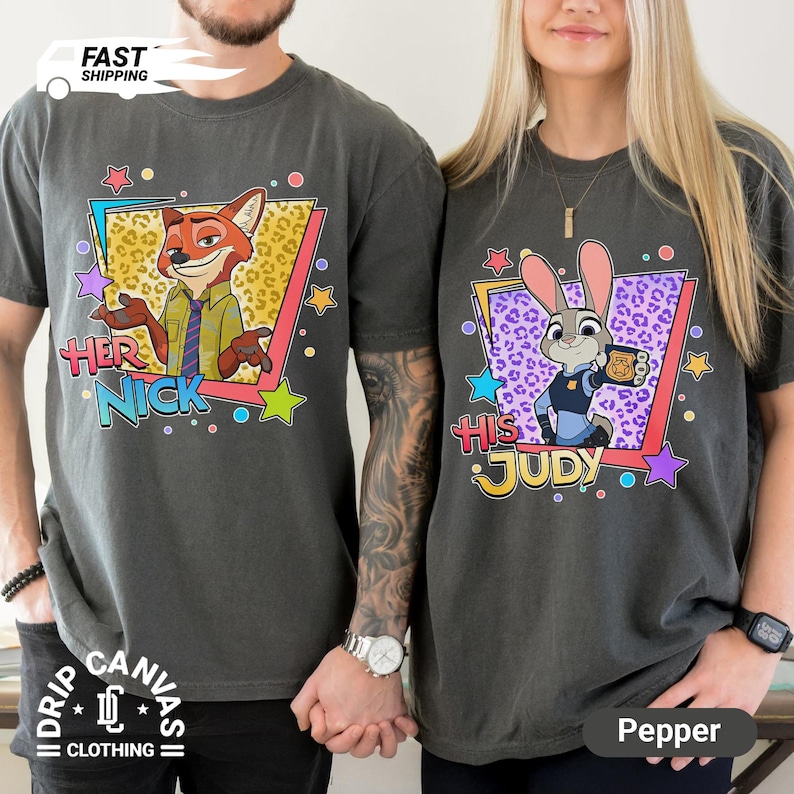 May include: Two dark gray t-shirts featuring characters from Zootopia. One shirt has Nick Wilde with "Her Nick" text, the other has Judy Hopps with "His Judy". Both shirts have colorful star and geometric designs.