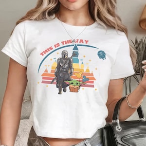 May include: White t-shirt featuring a graphic of a Mandalorian character and Grogu, with the text "THIS IS THE WAY". The design includes a colorful sunset and a spaceship. The shirt is a crew neck.