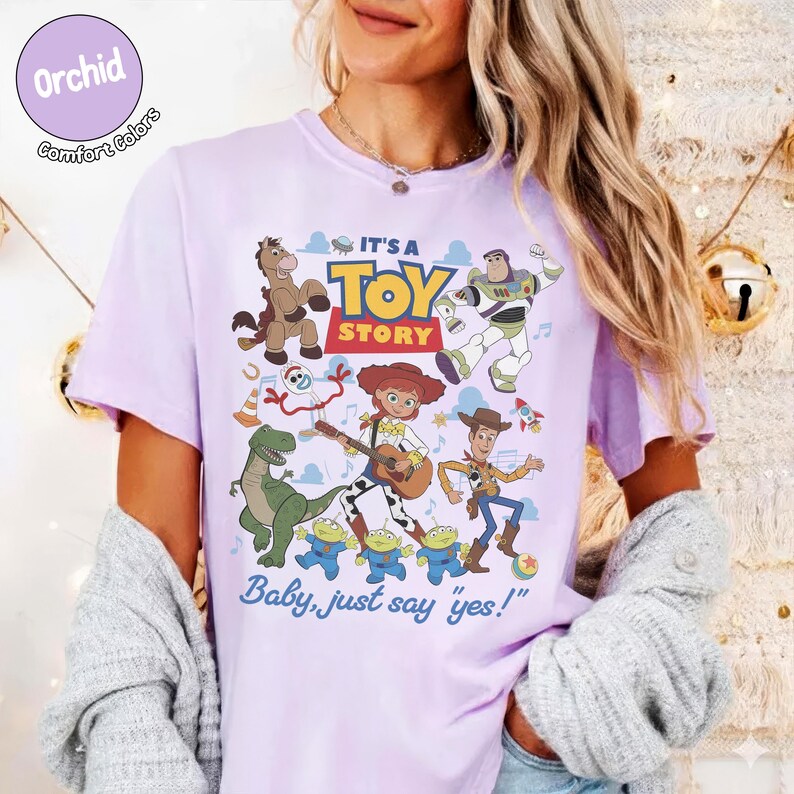 Op de afbeelding: Een lichtpaars T-shirt met korte mouwen en een grafische print van Toy Story-personages, waaronder Woody, Jessie, Buzz Lightyear, Rex, Bullseye, Forky en de aliens. De tekst luidt IT'S A TOY STORY en Baby, just say yes!