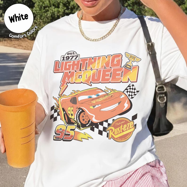 Op de afbeelding: Wit T-shirt met een afbeelding van Lightning McQueen uit de film Cars. Het ontwerp bevat de tekst "Lightning McQueen" en "95" met een geblokte vlag en het Rust-eze logo. Het shirt heeft een ronde hals.