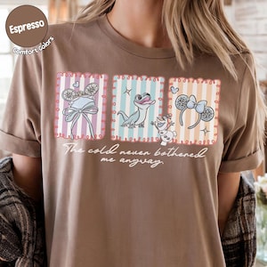 May include: A brown t-shirt with a graphic design featuring cartoon characters and accessories in framed images. The frames have striped pastel backgrounds. The text "The cold never bothered me anyway" is written below the images.