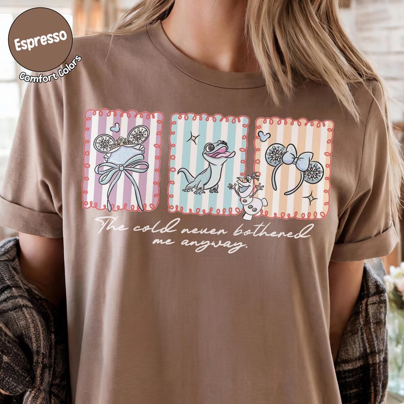 May include: A brown t-shirt with a graphic design featuring cartoon characters and accessories in framed images. The frames have striped pastel backgrounds. The text "The cold never bothered me anyway" is written below the images.