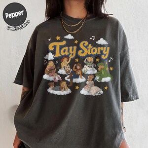 May include: A charcoal gray short-sleeve t-shirt featuring the text Tay Story in yellow with a blue outline. The graphic displays multiple illustrated figures resembling characters on clouds, surrounded by yellow stars, white clouds, and musical notes.